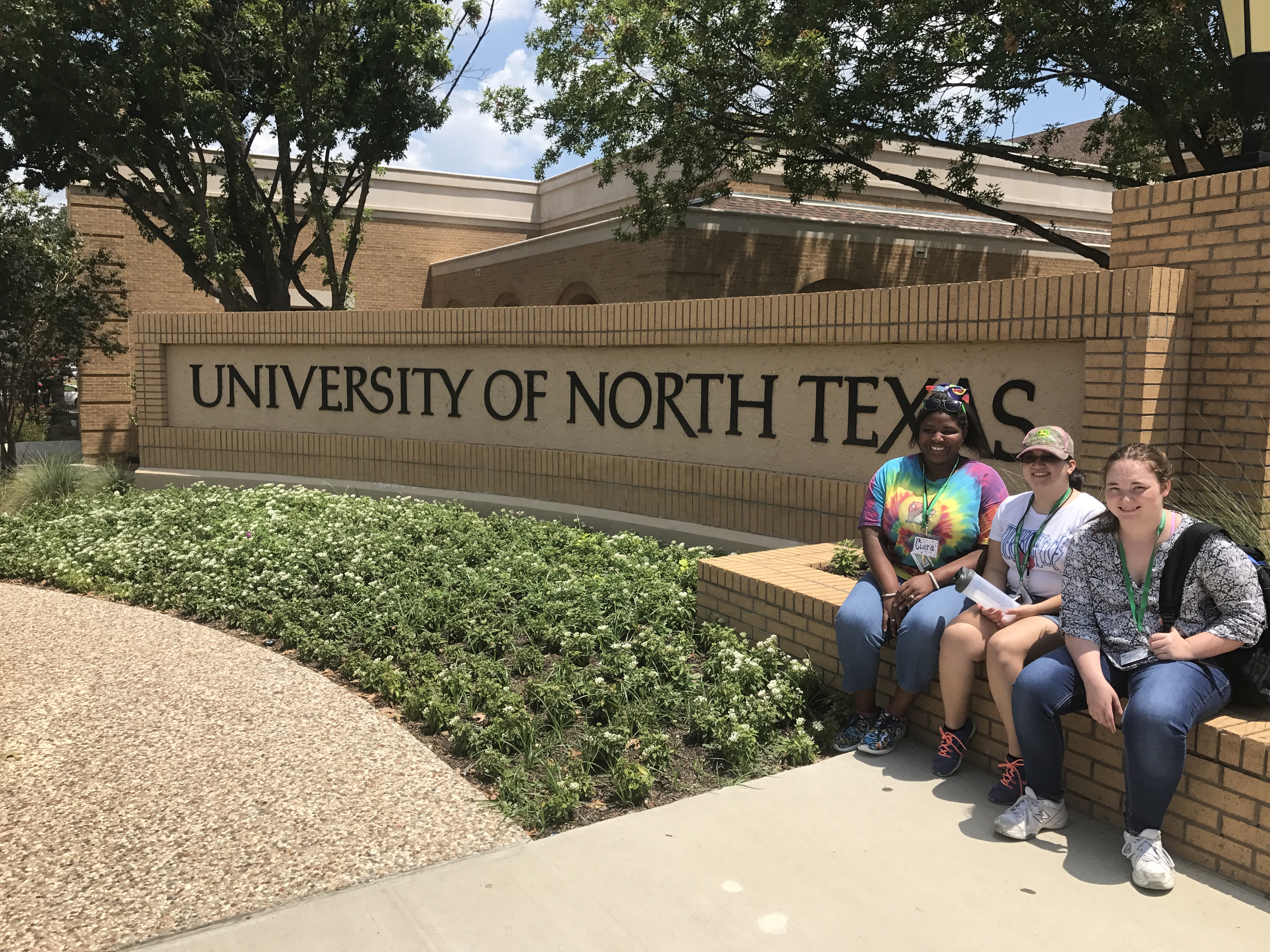 Three participants outside of Bruce Hall, sitting by a UNT sign. 